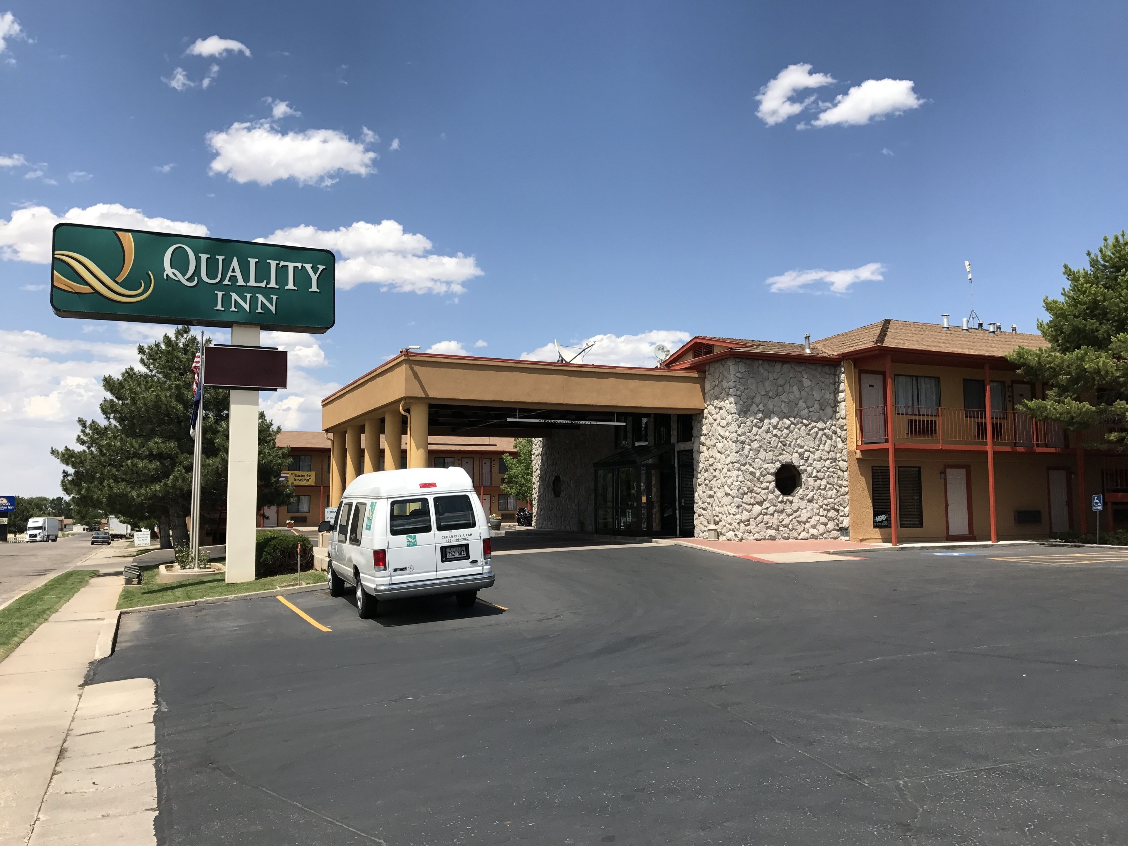 Un hôtel Quality Inn, reconnaissable à son enseigne bien visible, avec un fourgon garé devant et un bâtiment doté d’une façade en pierre et d’un balcon.
