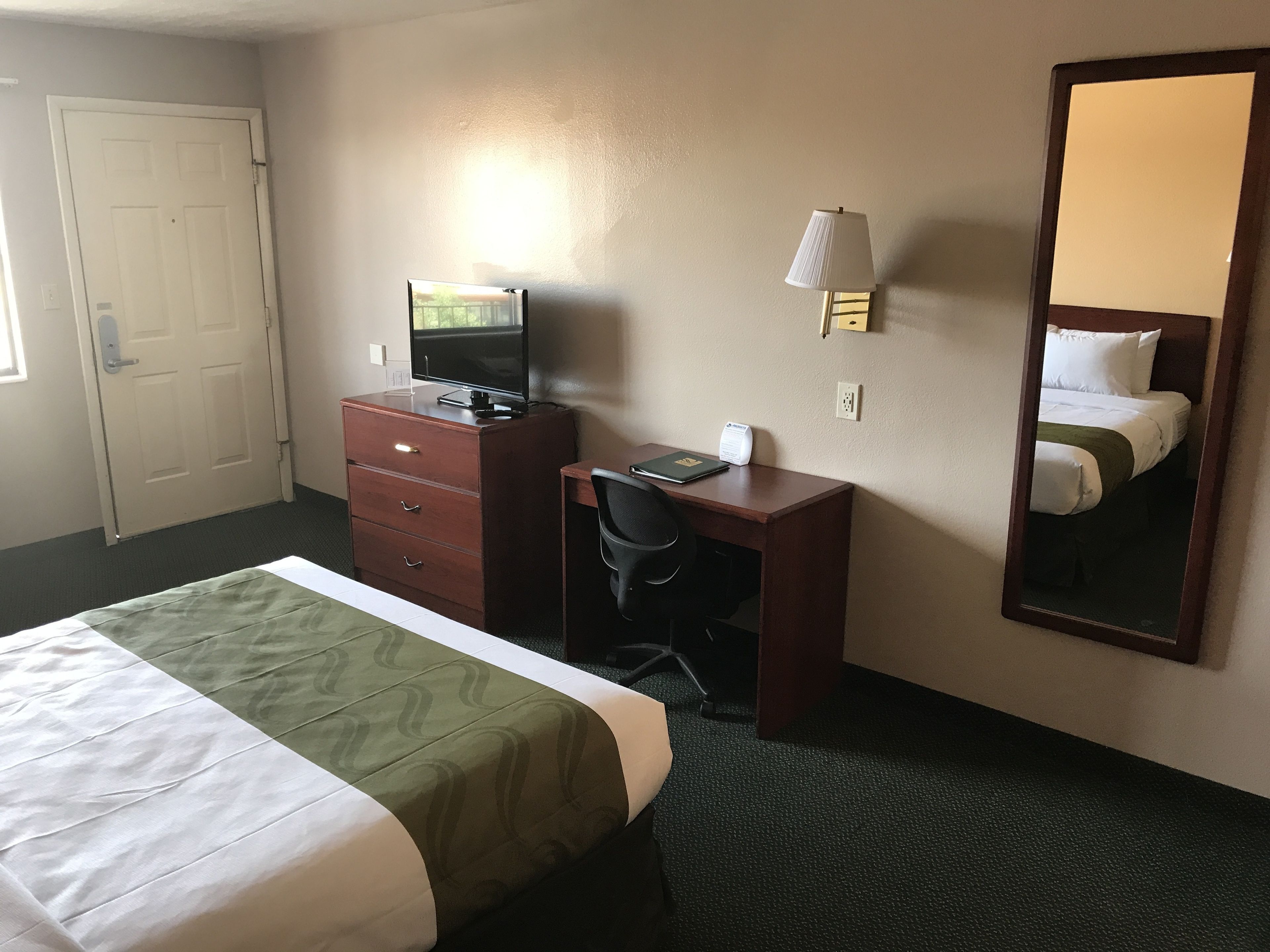A hotel room with a bed, a desk with a computer, a chair, a dresser, a lamp, and a mirror.