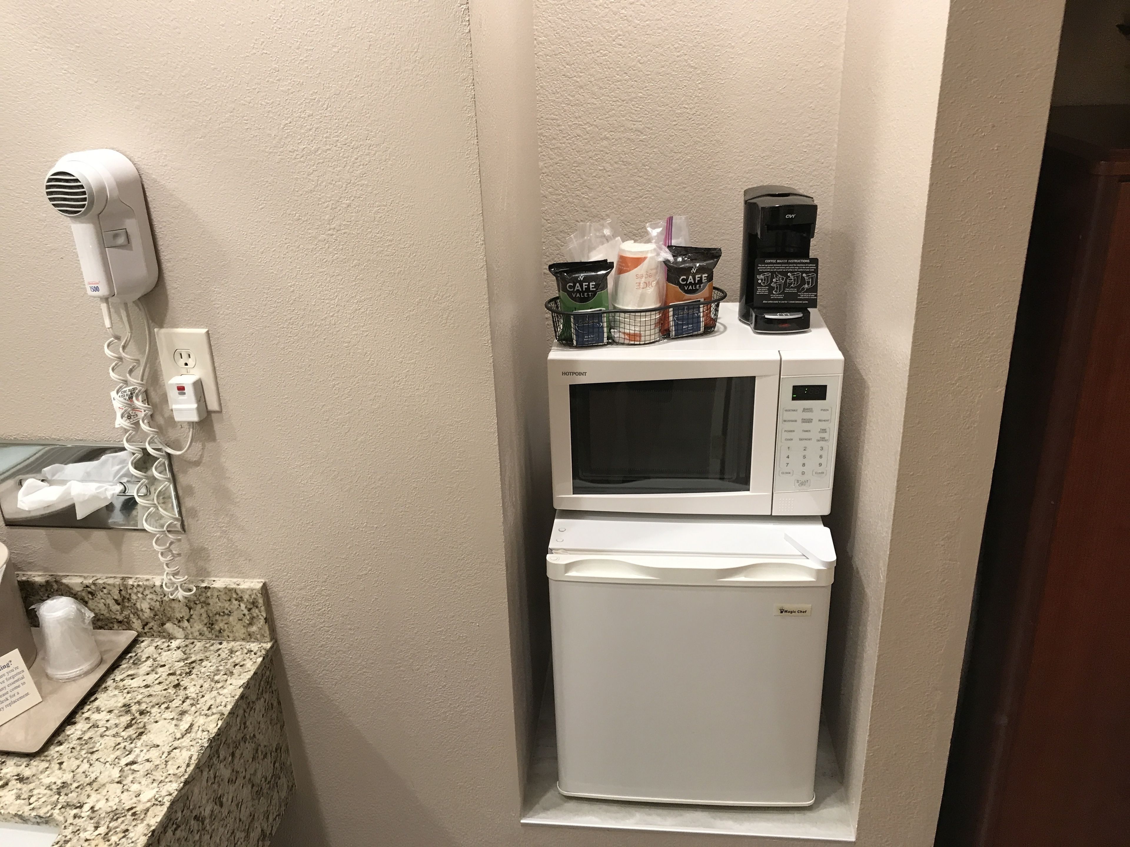 A compact kitchenette with a microwave, coffee maker, and a hair dryer plugged into a wall socket.