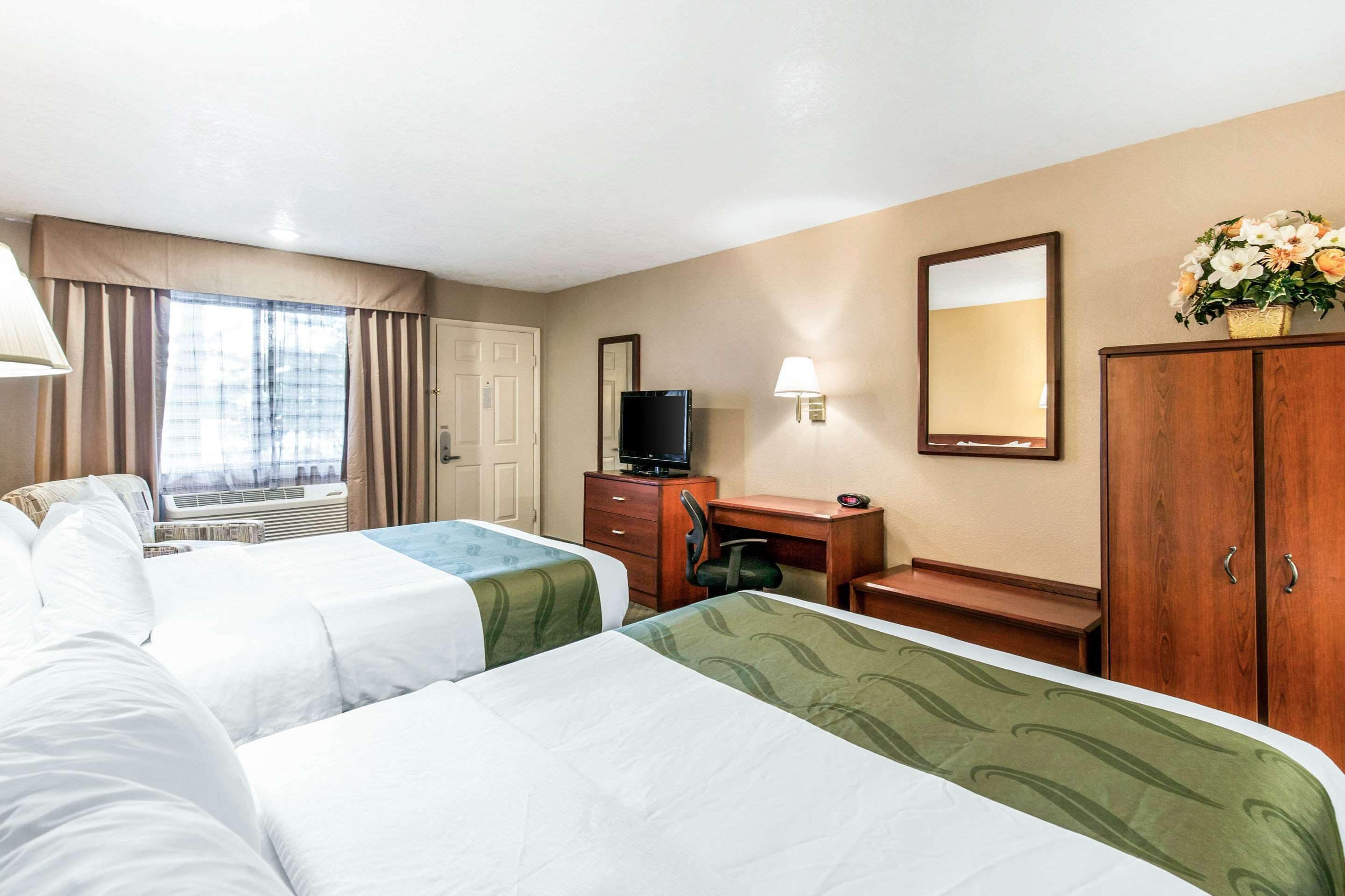 A hotel room with two beds, a desk, a television, a mirror, and a window with curtains.