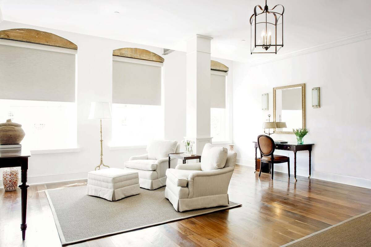 A spacious living room with a wooden floor, white armchairs, a small table, and a large mirror.
