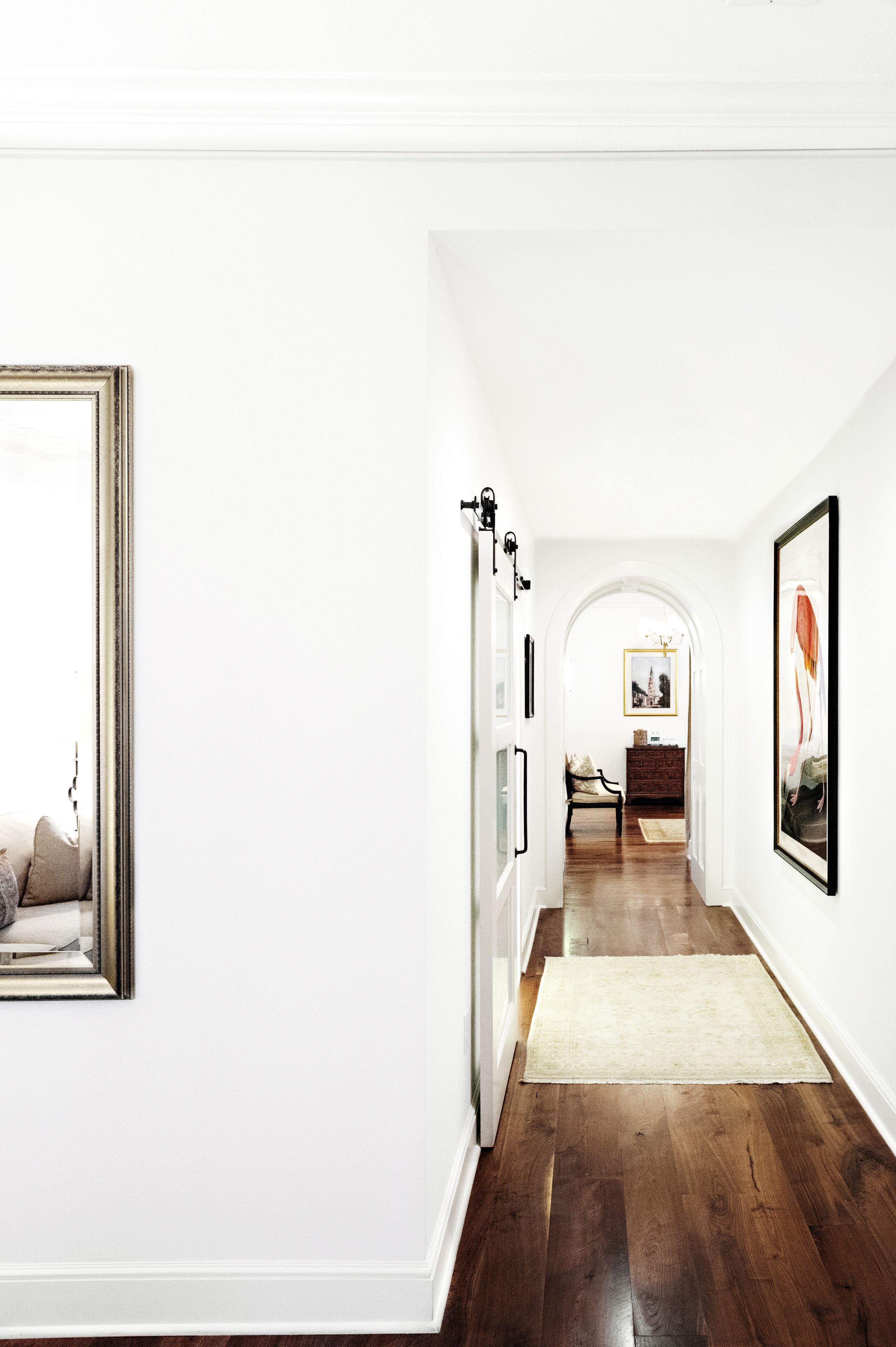 A hallway with a wooden floor, white walls, and a rug leading to a room with a chair and a dresser.
