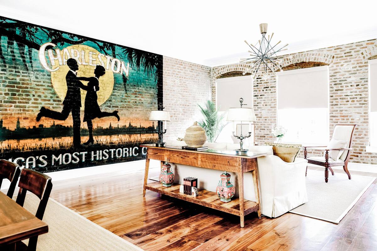 A room with a brick wall, a mural of Charleston, a wooden coffee table, and a white armchair.