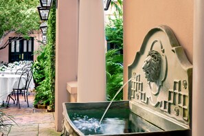 A fountain with a lion head sculpture, surrounded by greenery and a dining area with tables and chairs.