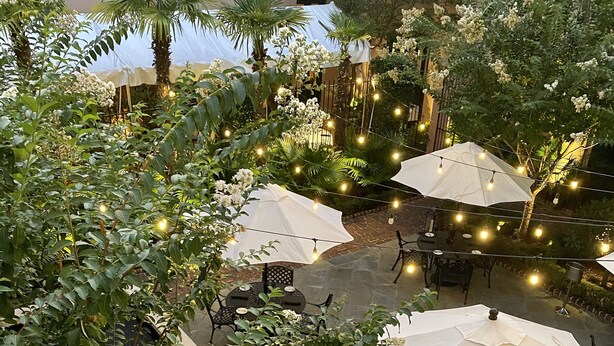 Un patio al aire libre con mesas y sombrillas, rodeado de exuberante follaje y una pequeña piscina.