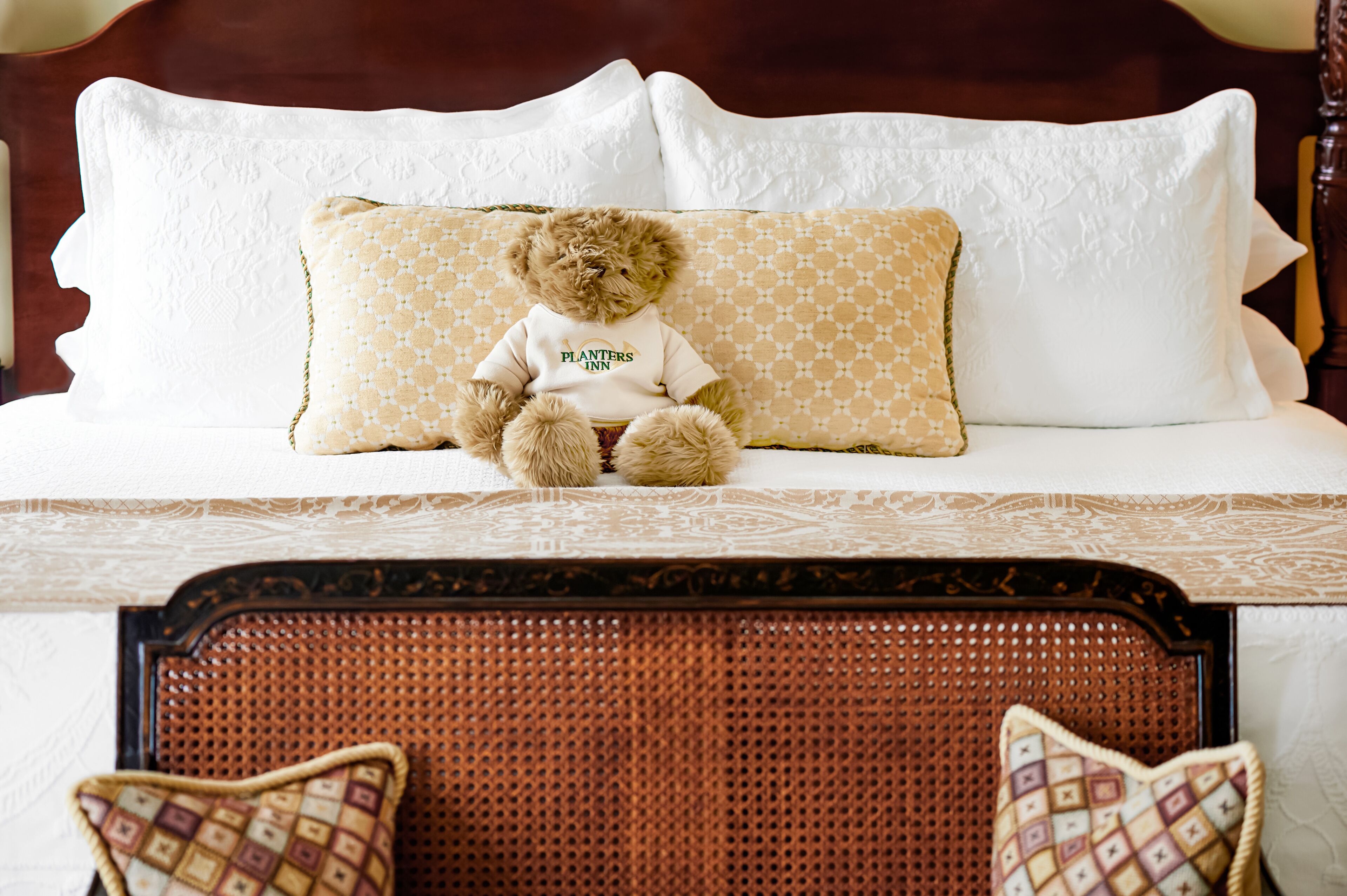 Una cama con una colcha blanca, un osito de peluche marrón que lleva una camisa blanca y almohadas estampadas.