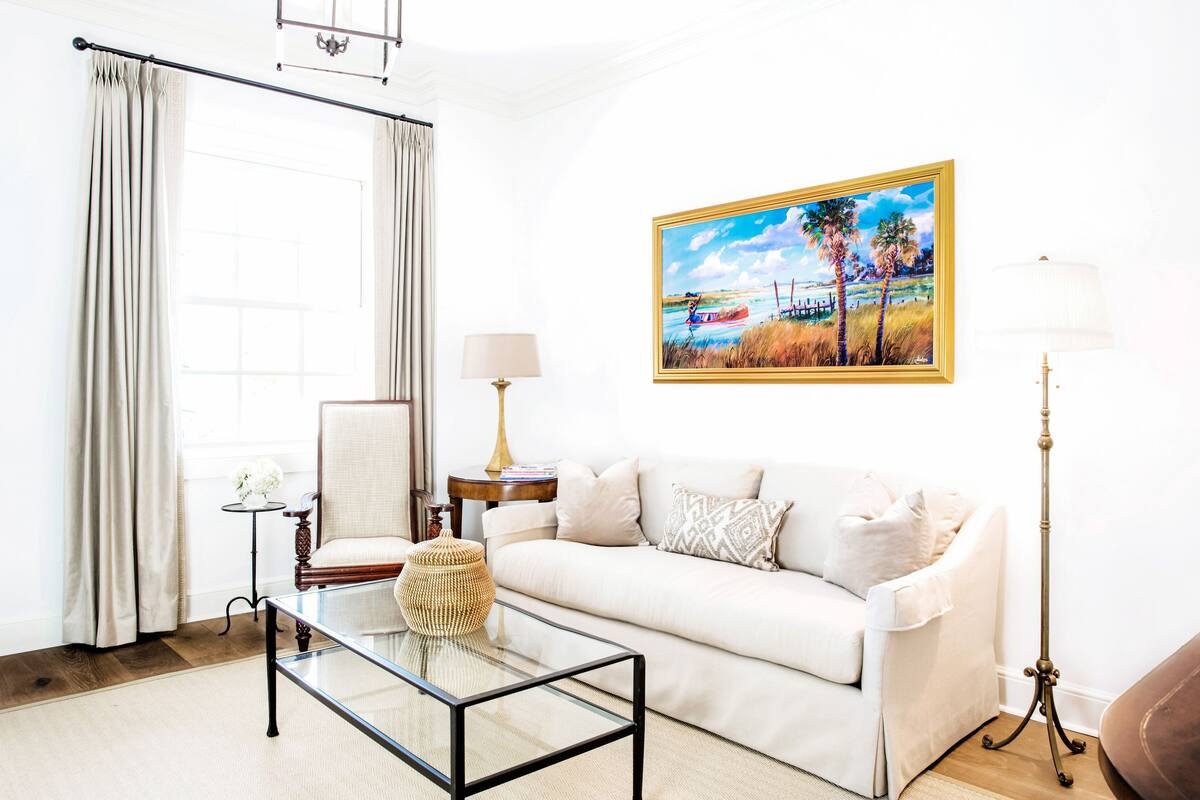 A living room with a white sofa, a glass coffee table, a side table with a lamp, a painting on the wall, and a view of the outdoors through a window with curtains.