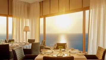 Un comedor con una mesa dispuesta para dos, ventanas amplias con cortinas y vistas al mar.