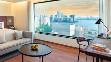 Une chambre d’hôtel moderne avec une grande fenêtre, un canapé, une table ronde avec des fruits, un bureau avec des livres et une chaise.