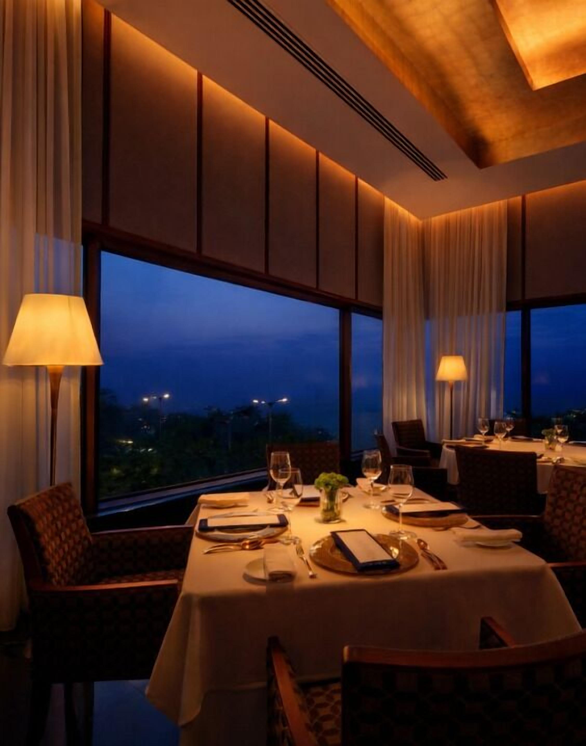 A dining area with a table set for two, large windows, and a view of the cityscape at dusk.