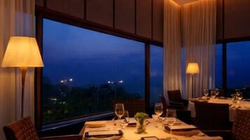A dining area with a table set for two, large windows, and a view of the cityscape at dusk.