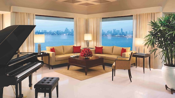 A spacious living room with a grand piano, a sofa set, a coffee table, and a large window offering a city skyline view. - The Oberoi Mumbai (Mumbai)
