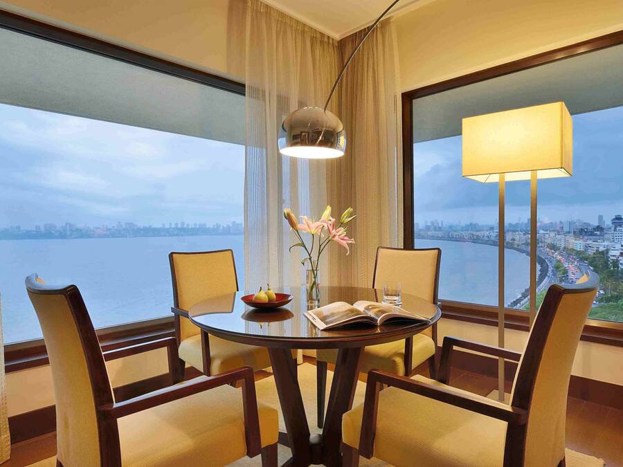 A hotel room with a round table, four chairs, a vase with flowers, a bowl of fruit, and a book.