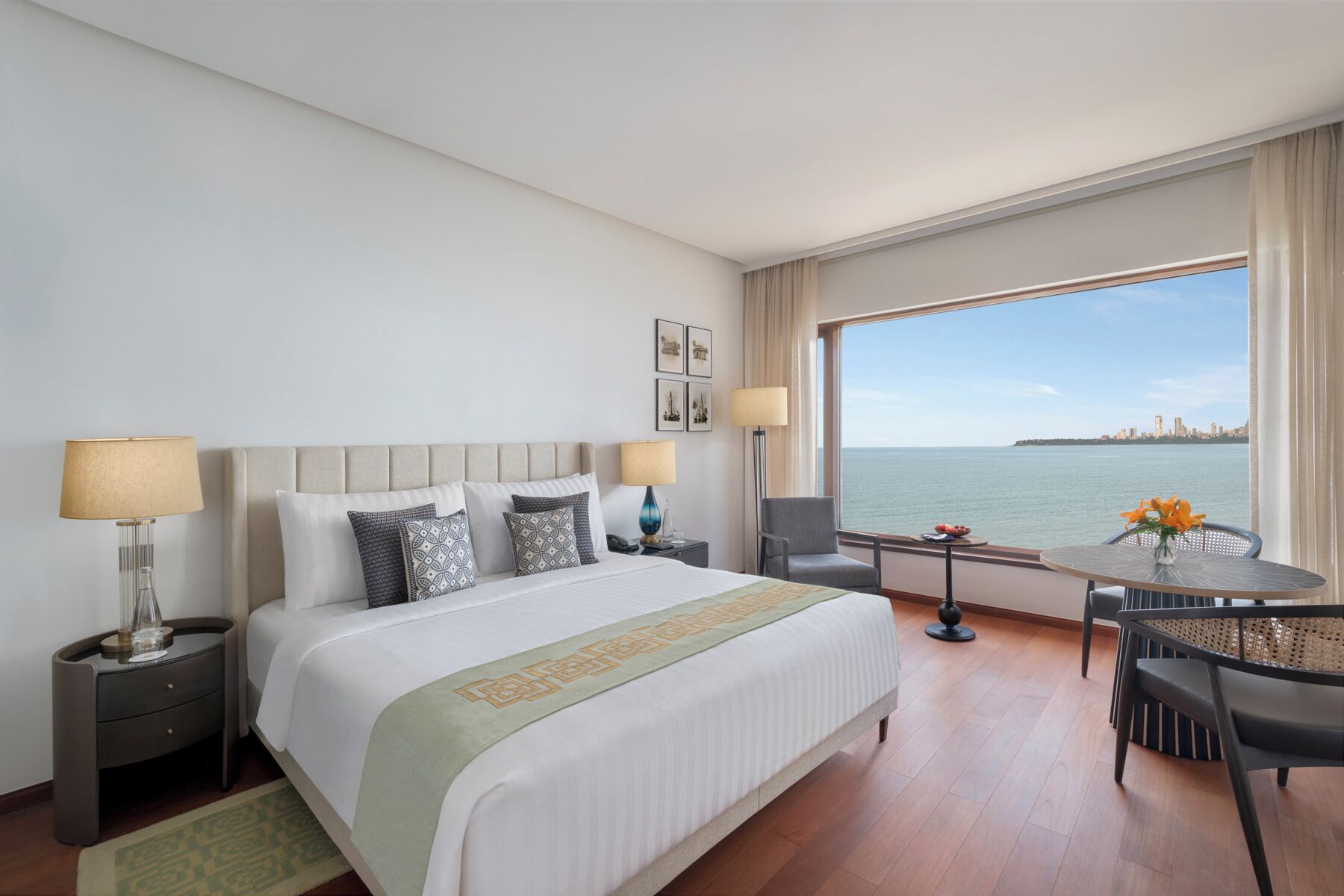 Habitación de hotel con una cama, dos mesitas de noche, una silla, una pequeña mesa y vistas al mar.