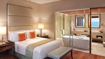 A modern hotel room with a large bed, bedside lamps, a bench, a glass-enclosed bathroom with a bathtub, and a view of the sea.