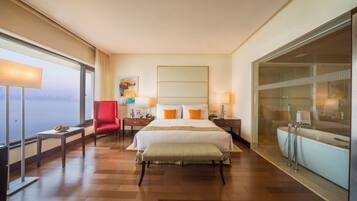 Quarto de hotel com uma cama grande, uma cadeira vermelha, mesa de cabeceira e banheiro com vidro, além de vista para a cidade.