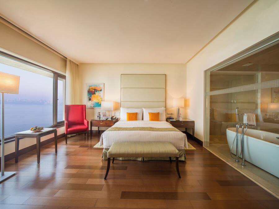 Quarto de hotel com uma cama grande, uma cadeira vermelha, mesa de cabeceira e banheiro com vidro, além de vista para a cidade.
