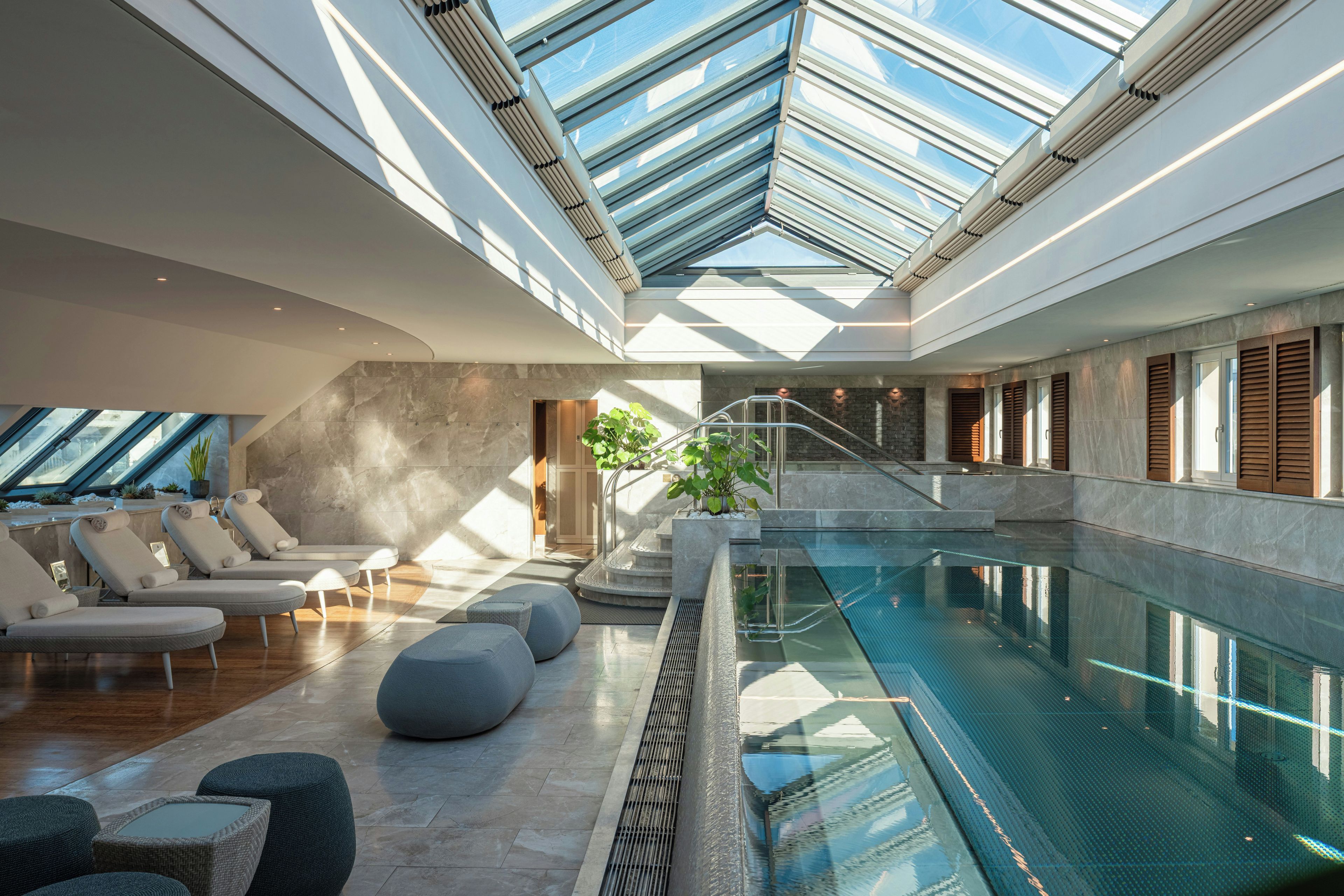 A modern indoor swimming pool with a glass ceiling, lounge chairs, and a staircase leading to a lower level.