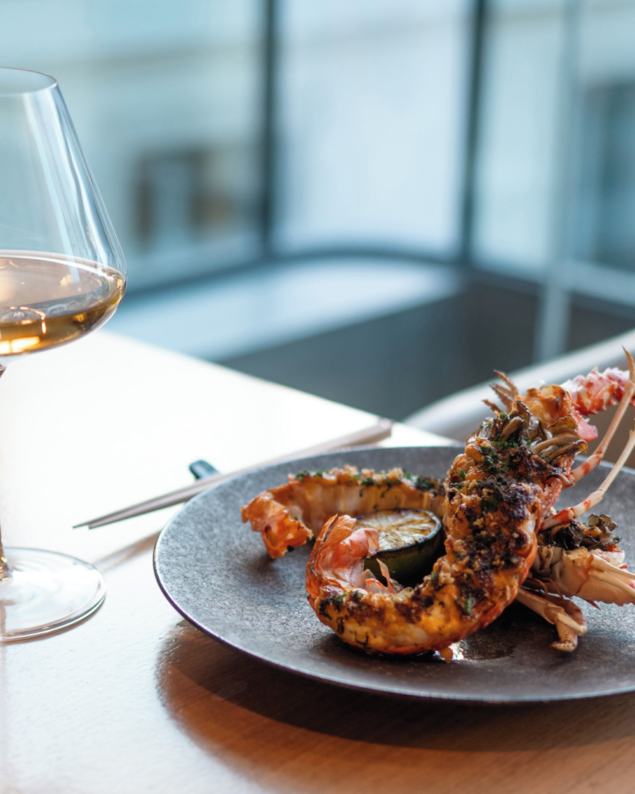 Une assiette noire sur laquelle repose un homard cuit, un verre de vin blanc et un stylo posés sur une table.