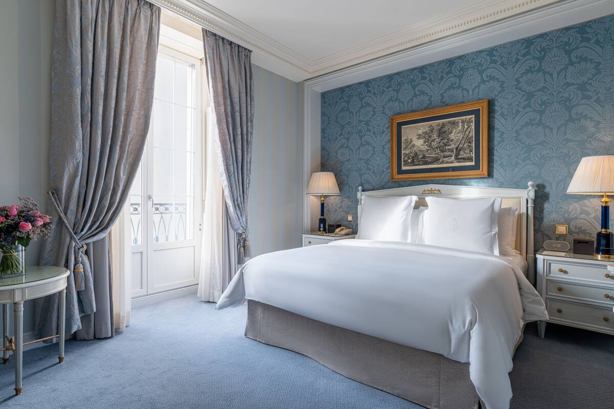 Une chambre à coucher avec un grand lit, deux tables de chevet, une commode, un tableau encadré au mur et une fenêtre avec des rideaux.