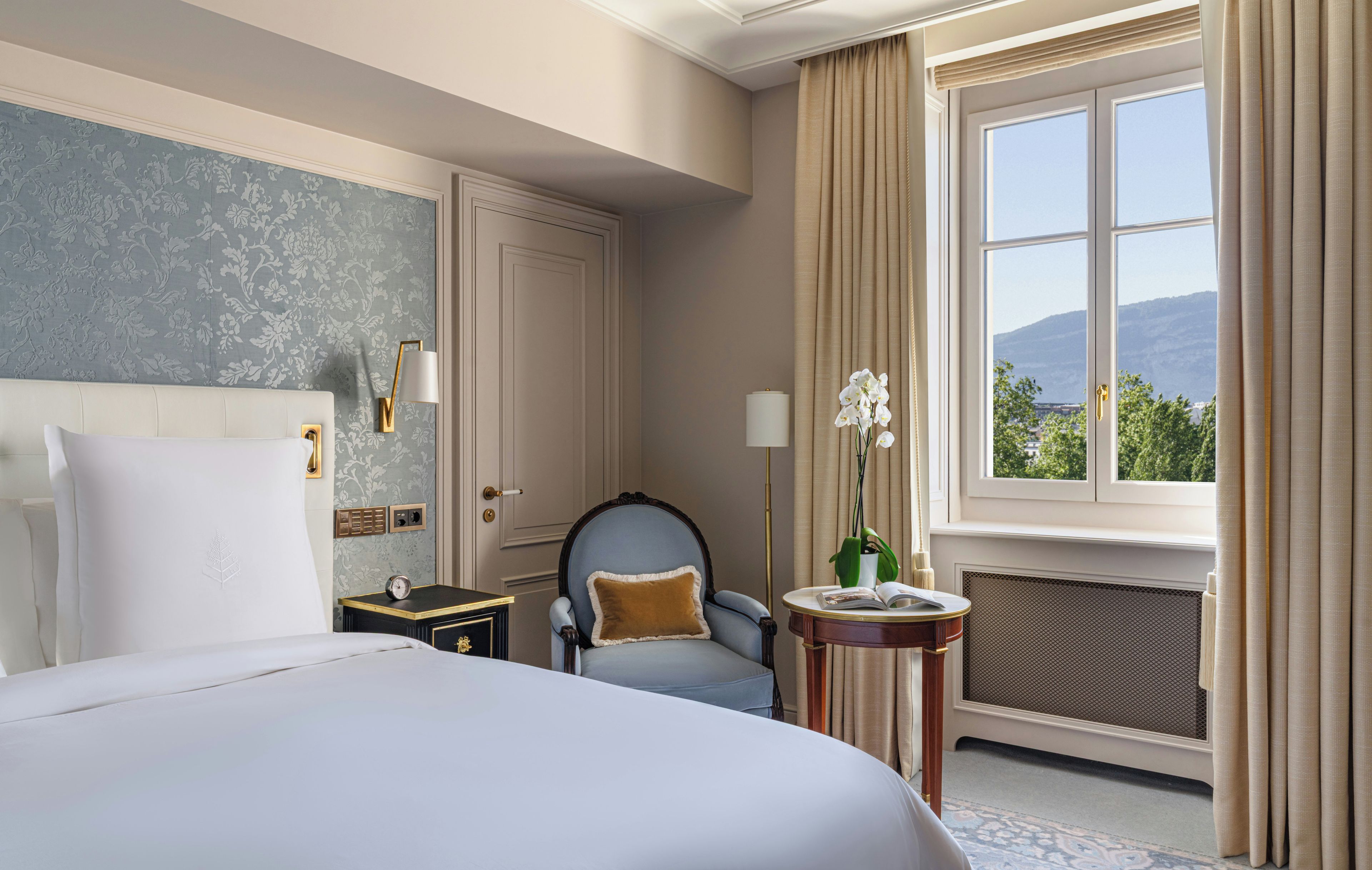 Een hotelkamer met een bed, een stoel, een klein tafeltje met een plant, een raam met gordijnen en een radiator.