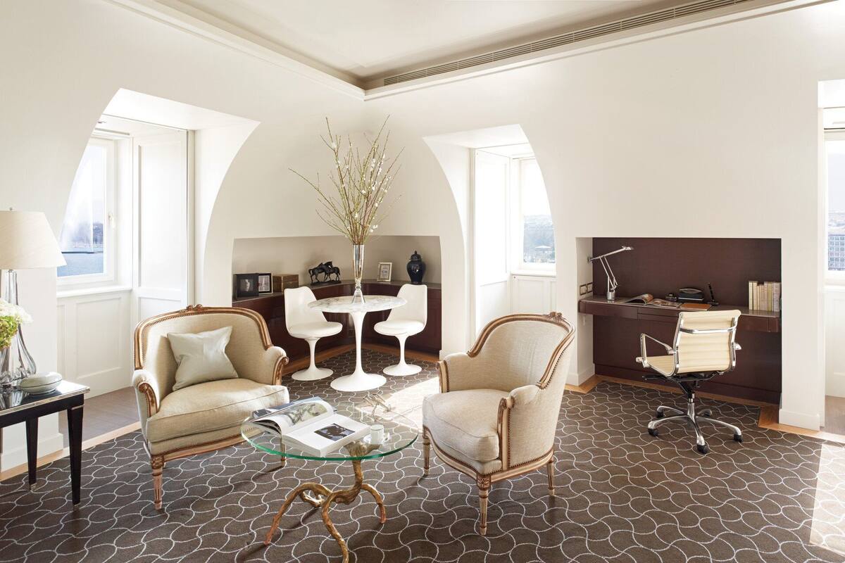 Un salon moderne avec une table basse en verre, deux fauteuils, un bureau avec un ordinateur et une vue sur le paysage urbain, visible à travers de grandes fenêtres.