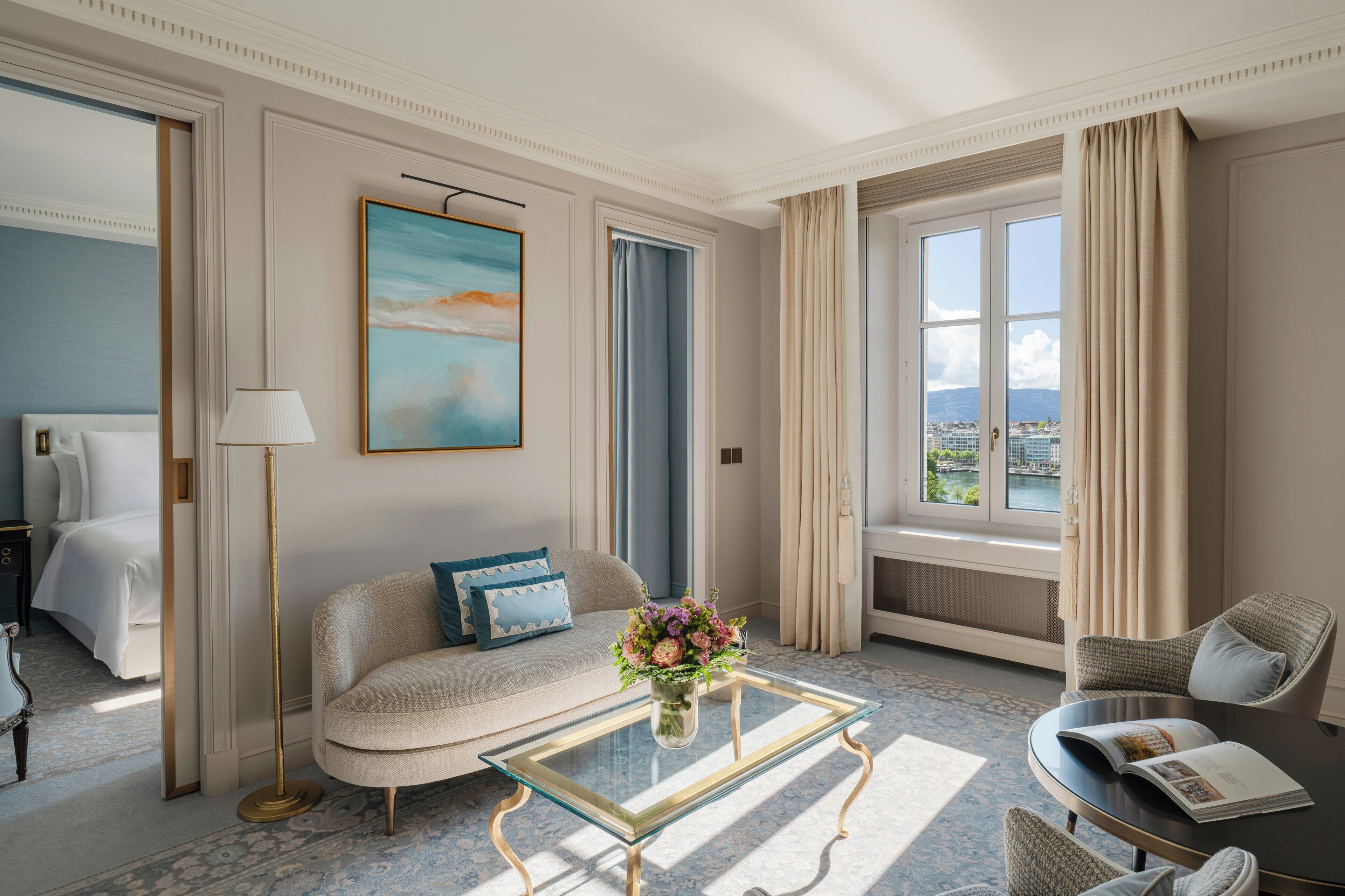 Una habitación de hotel con cama, sofá, sillón, una mesa de centro de vidrio con un jarrón de flores, un cuadro en la pared y vistas de la ciudad a través de la ventana.