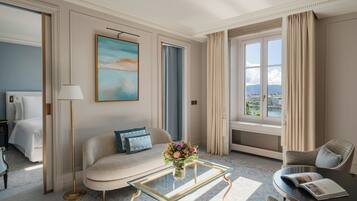 Una habitación de hotel con cama, sofá, sillón, una mesa de centro de vidrio con un jarrón de flores, un cuadro en la pared y vistas de la ciudad a través de la ventana.