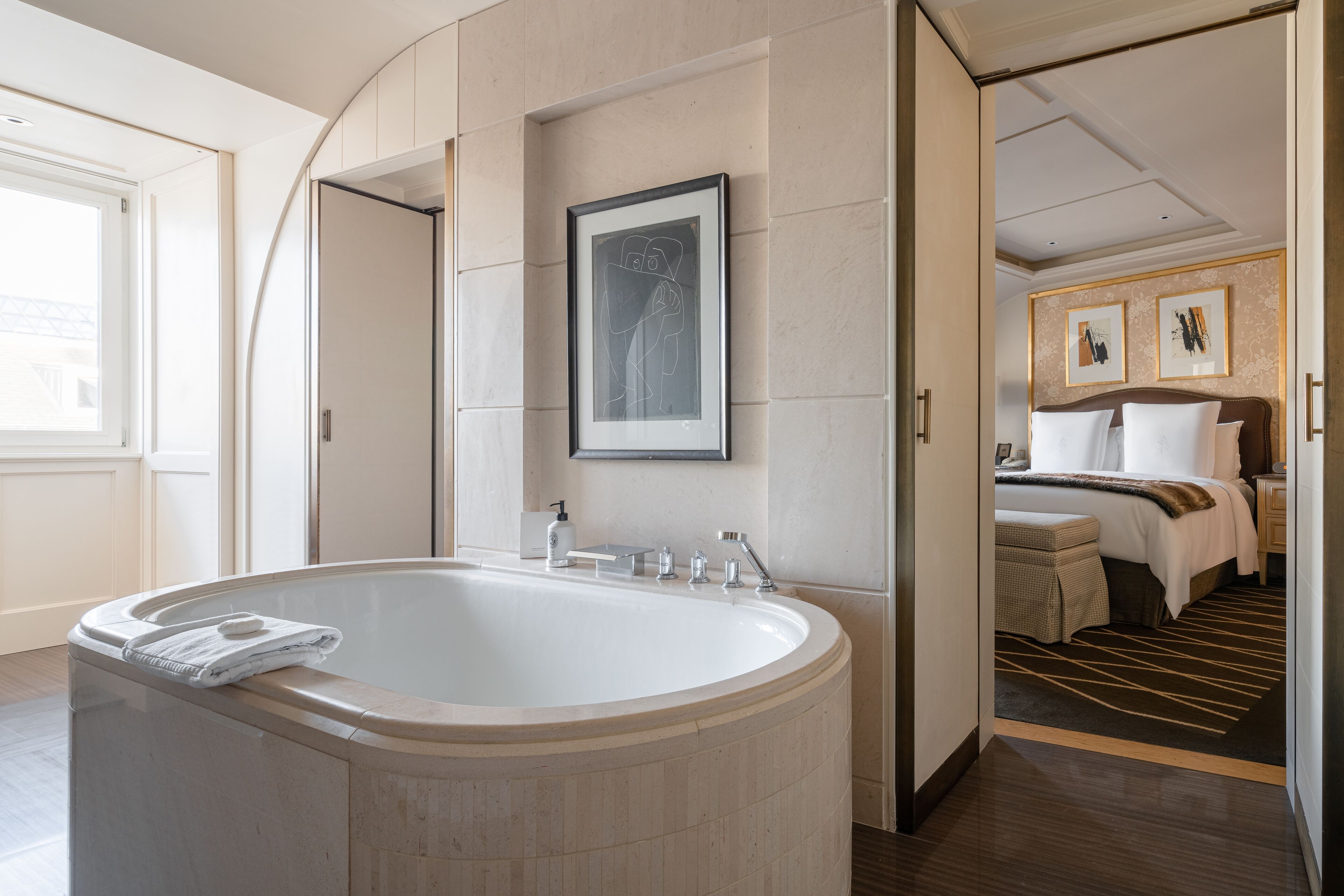 Een moderne badkamer met een vrijstaand bad, een kunstwerk in een lijst aan de muur, en een zichtbare slaapkamer met een bed en nachtkastje.