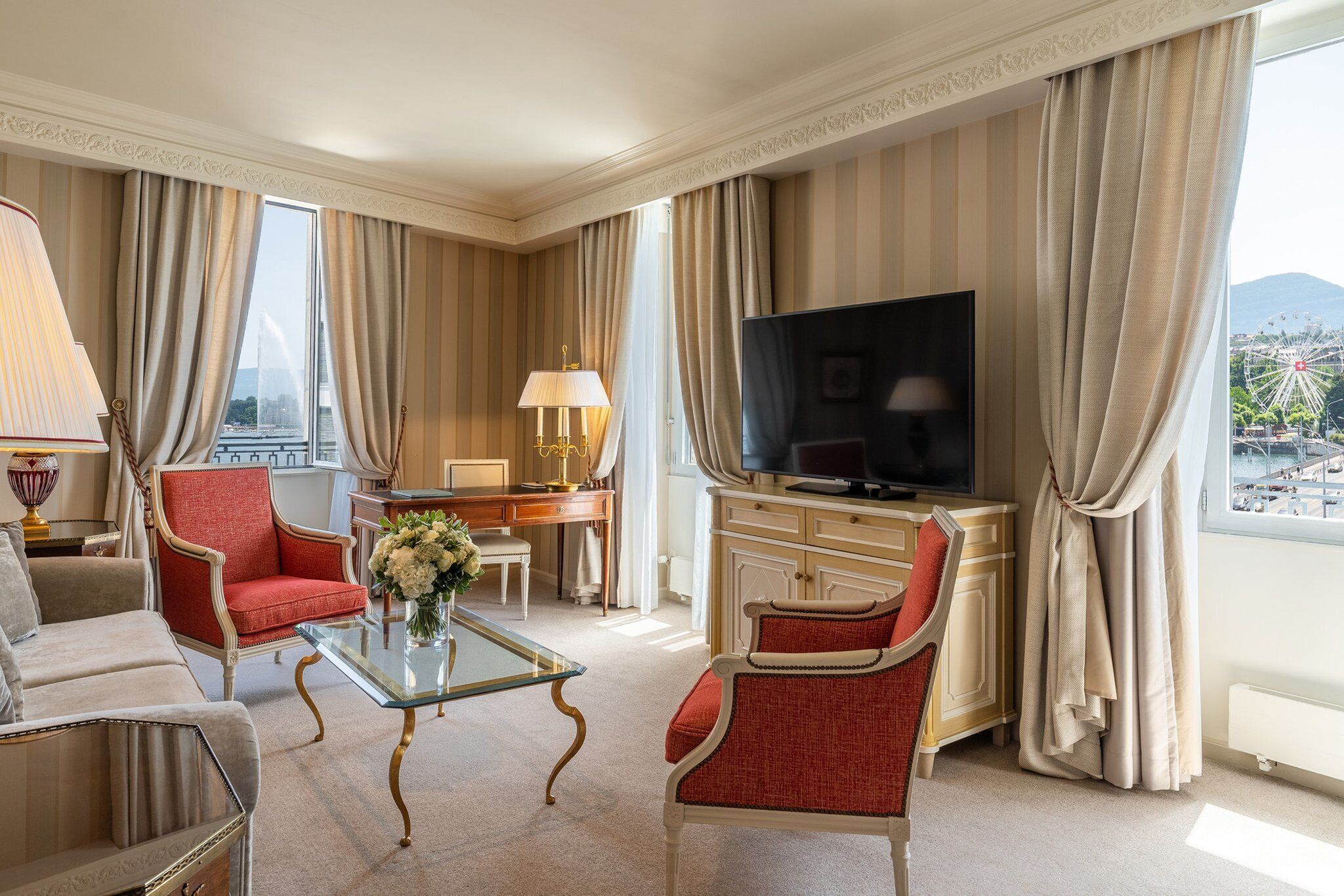 Una habitación de hotel espaciosa con un televisor de pantalla plana grande, una zona de estar confortable con sillones tapizados en rojo, una mesa de centro de vidrio y vistas a una noria a través de amplios ventanales.