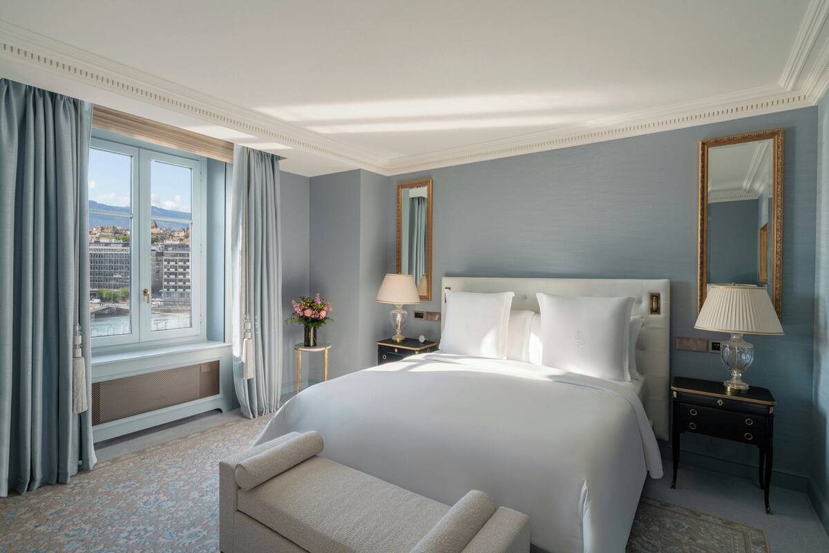 Une chambre d’hôtel avec un grand lit, un banc, une fenêtre avec des rideaux, une table de chevet avec une lampe et un miroir.