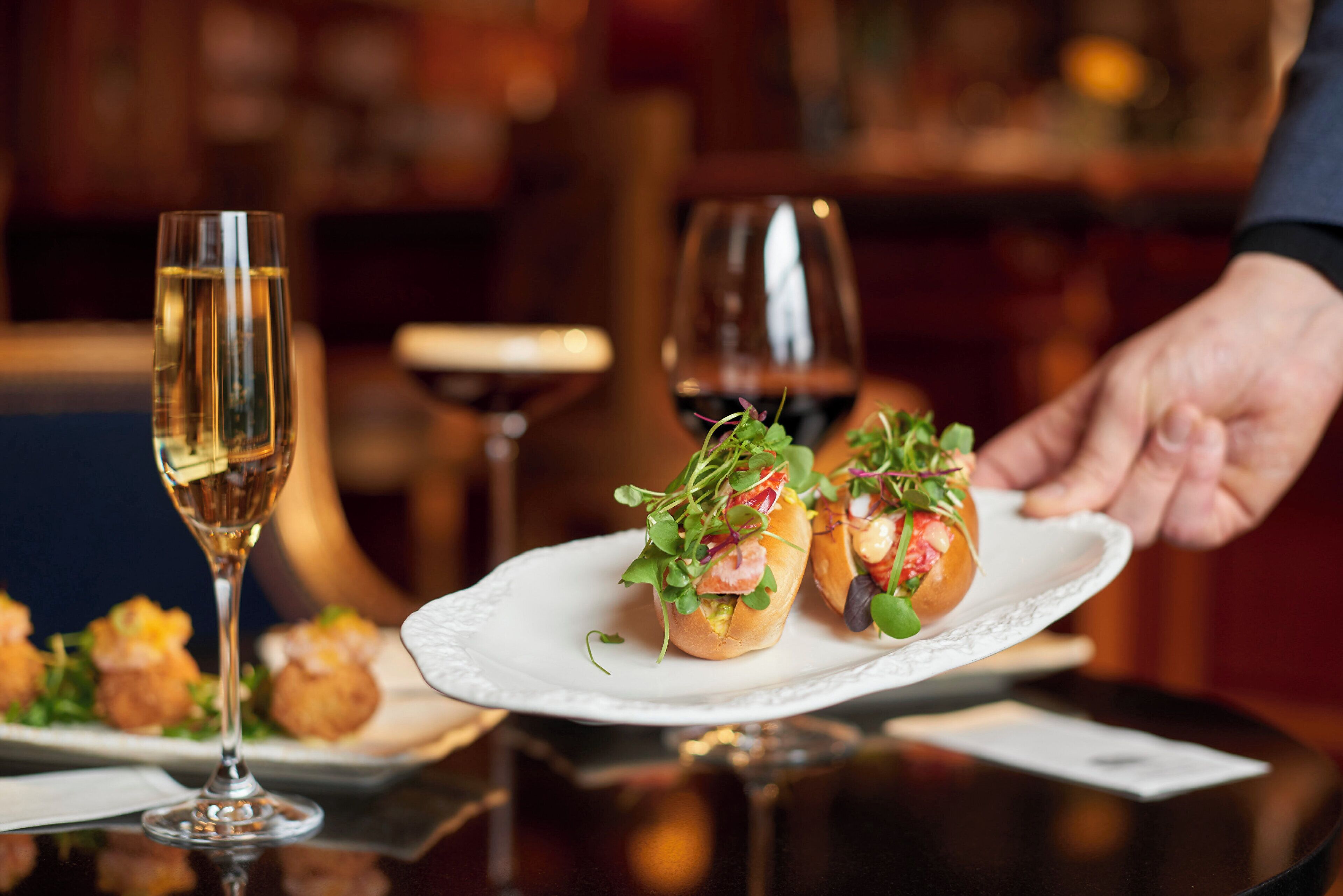 A dining table with a white plate holding two appetizers, a glass of white wine, and a wine glass.