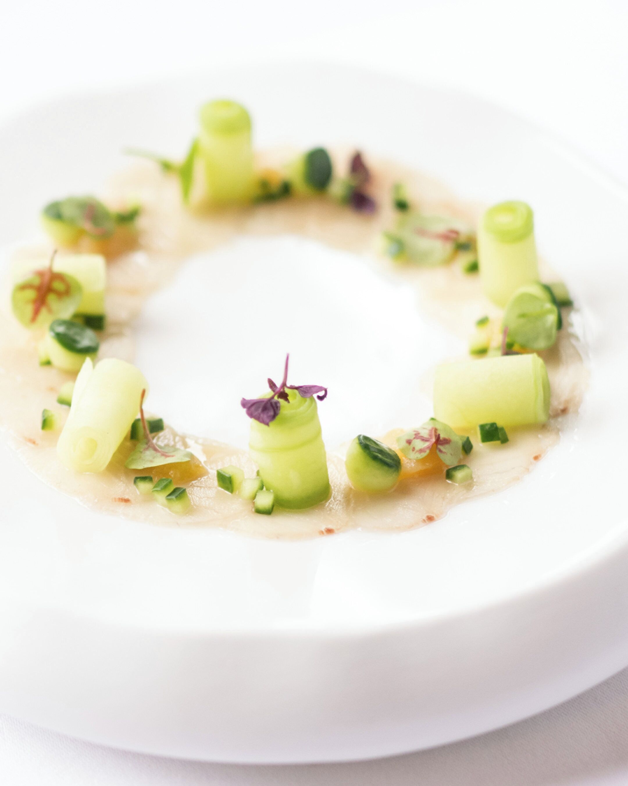 A white plate with a circular arrangement of food items, including green vegetables and possibly some meat, garnished with small purple flowers.