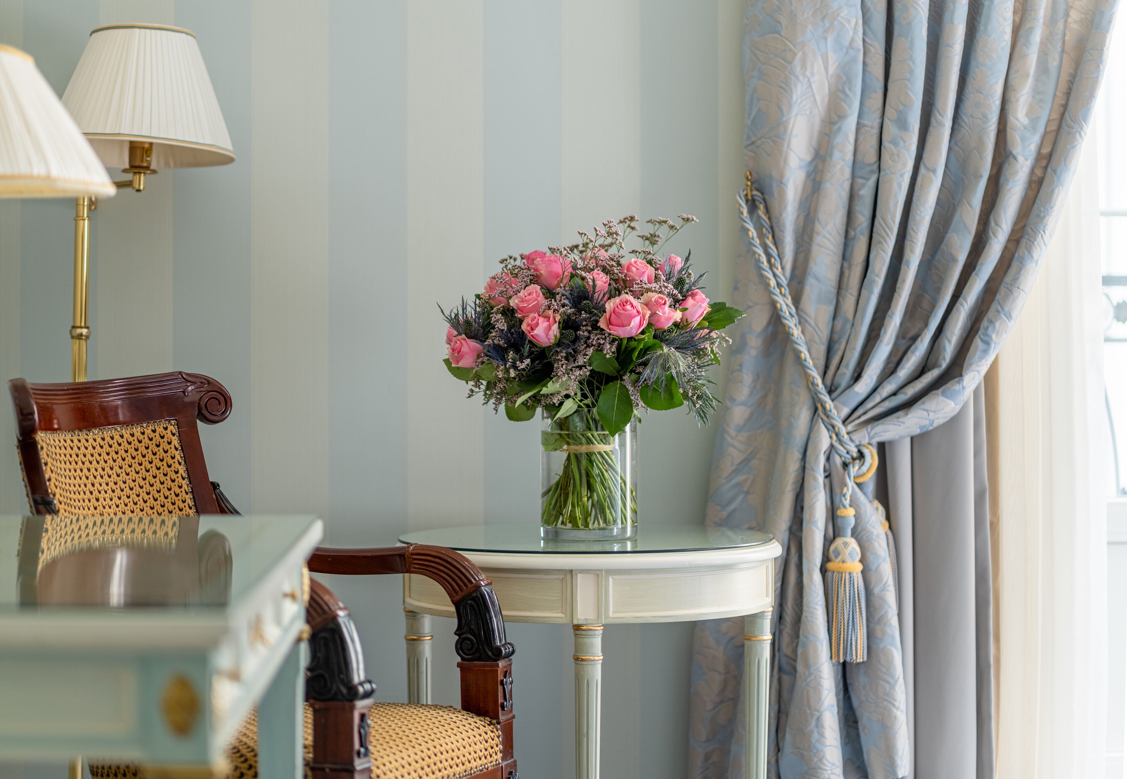 A room with a round glass table, a vase of pink flowers, a lamp, and a chair with a woven seat.