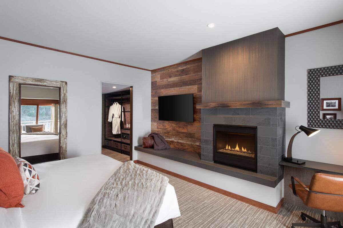 A modern hotel room with a large bed, a fireplace, a mounted TV, and a view of the outdoors.