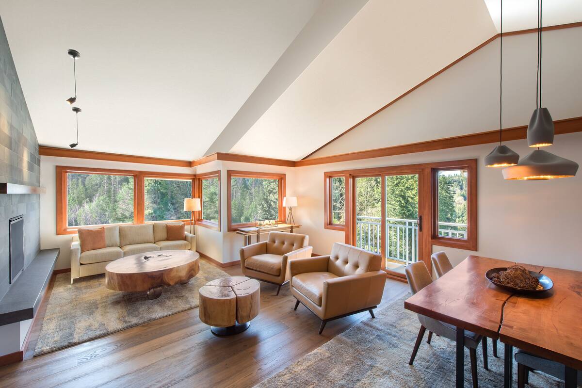 A spacious living room with a large wooden dining table, a sofa, and two armchairs. There are large windows providing natural light and a view of the outdoors.