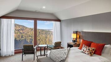 A hotel room with a large bed, a desk, two chairs, a view of a forested area, and a window with curtains.