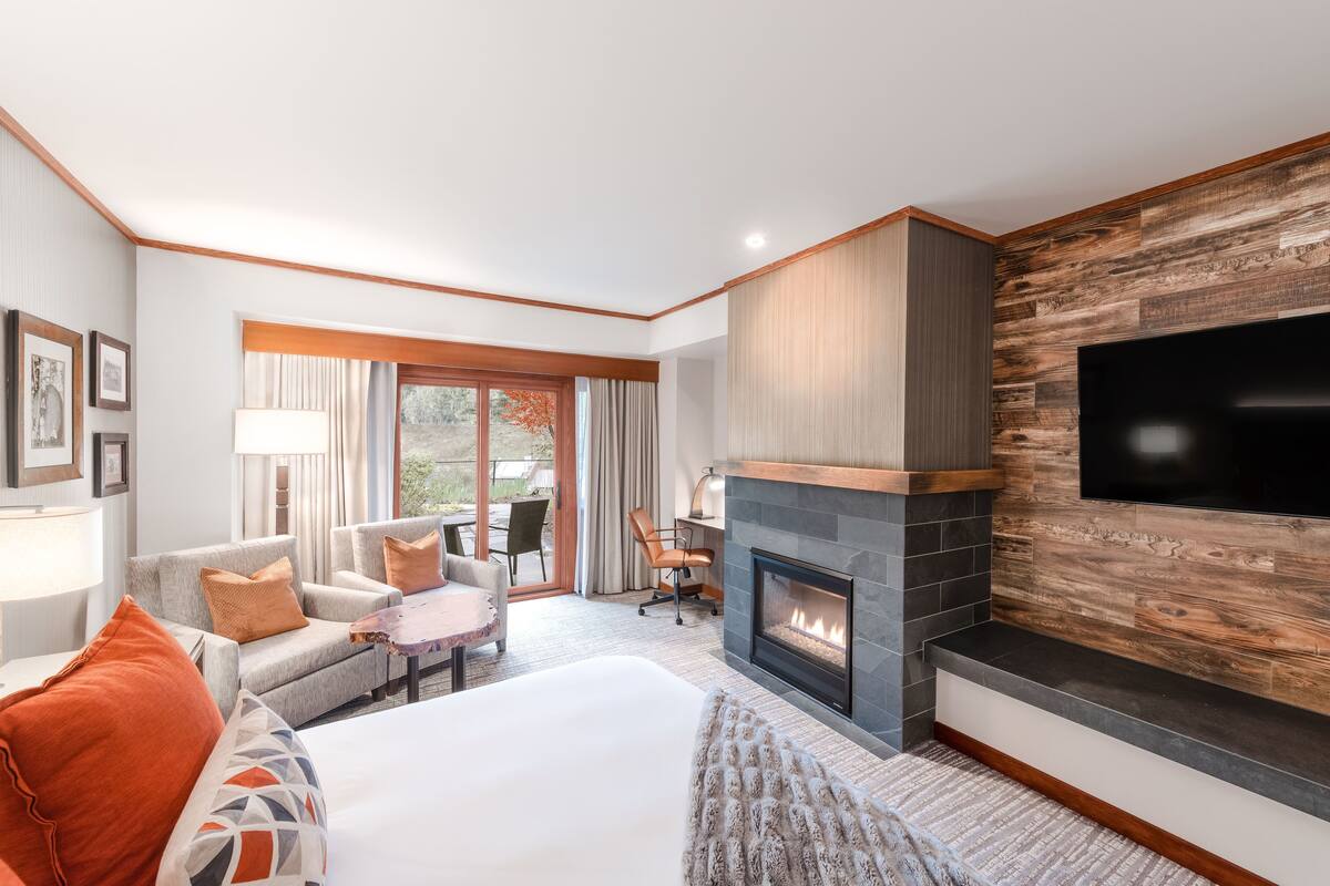 A modern hotel room with a large bed, a fireplace, a TV mounted on the wall, and a seating area with a desk and chairs.