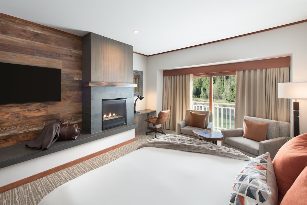 A modern hotel room with a fireplace, a large bed, a seating area with a view, and a flat-screen TV mounted on the wall.