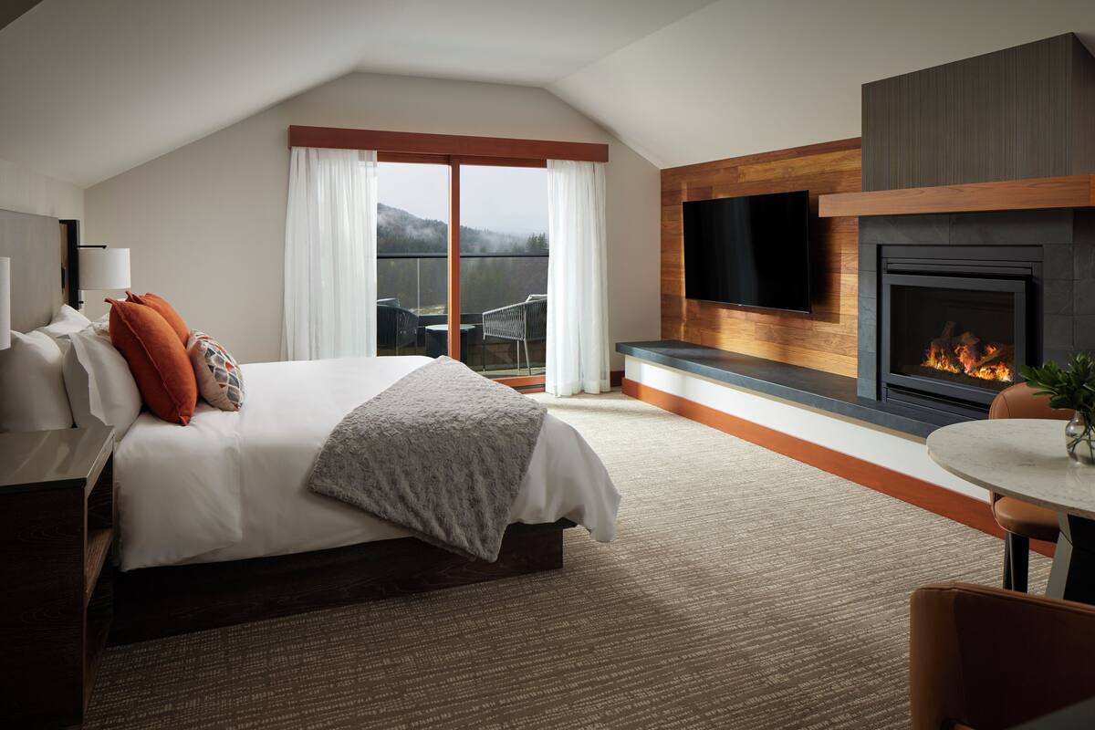 A modern hotel room with a large bed, a fireplace, a TV, and a balcony with a view.