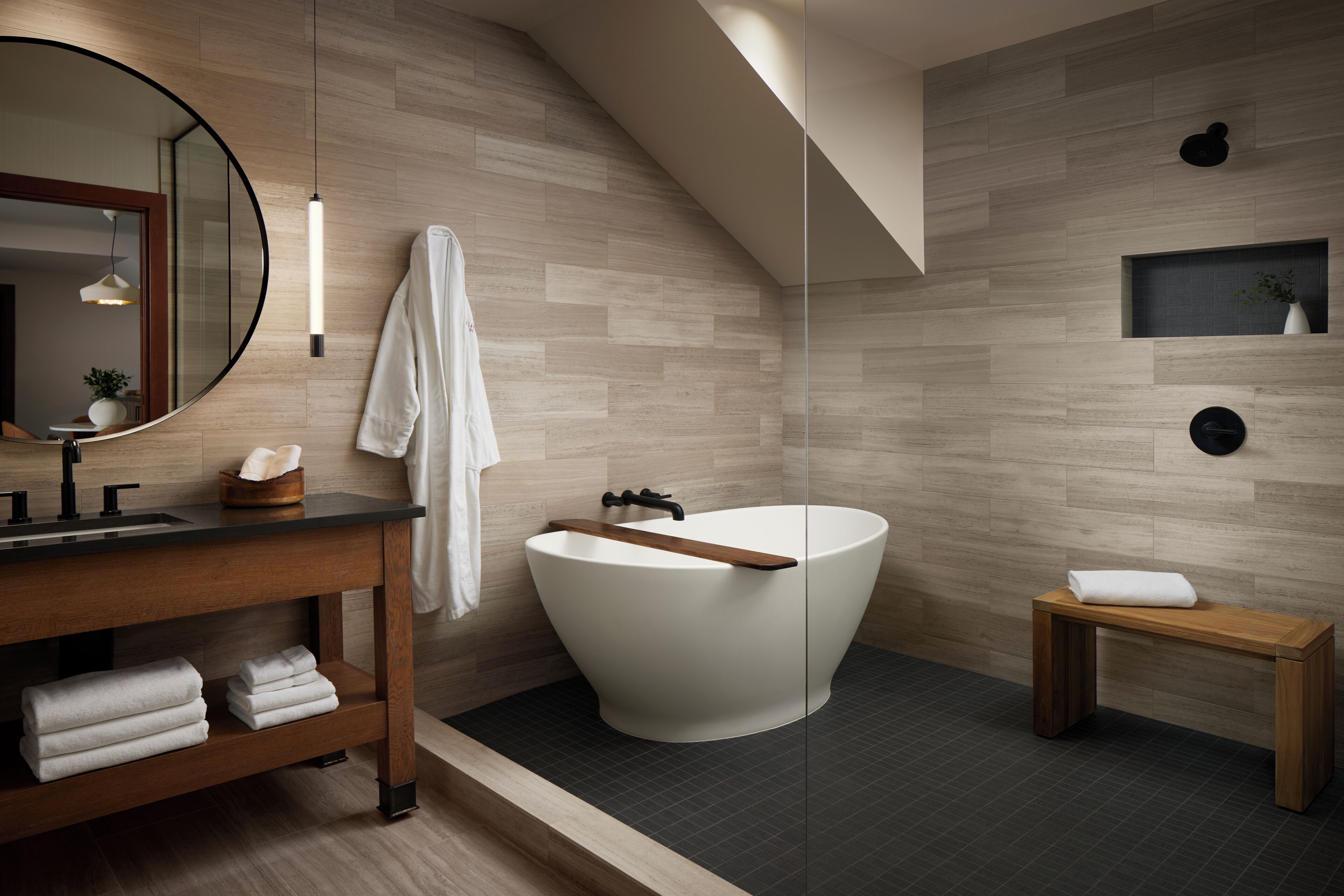 A modern bathroom with a freestanding bathtub, a wooden vanity, a round mirror, and a small shelf with a plant.