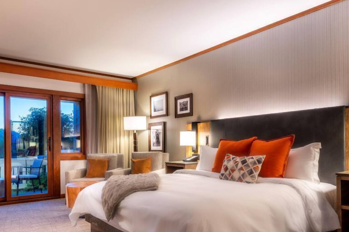 A hotel room with a large bed, a desk, a chair, and a view of the outdoors through a sliding door.