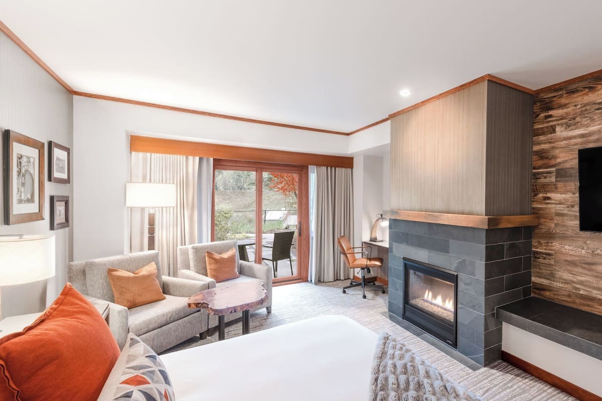A modern hotel room with a fireplace, a large flat-screen TV, a sofa, a desk, and a view of the outdoors.
