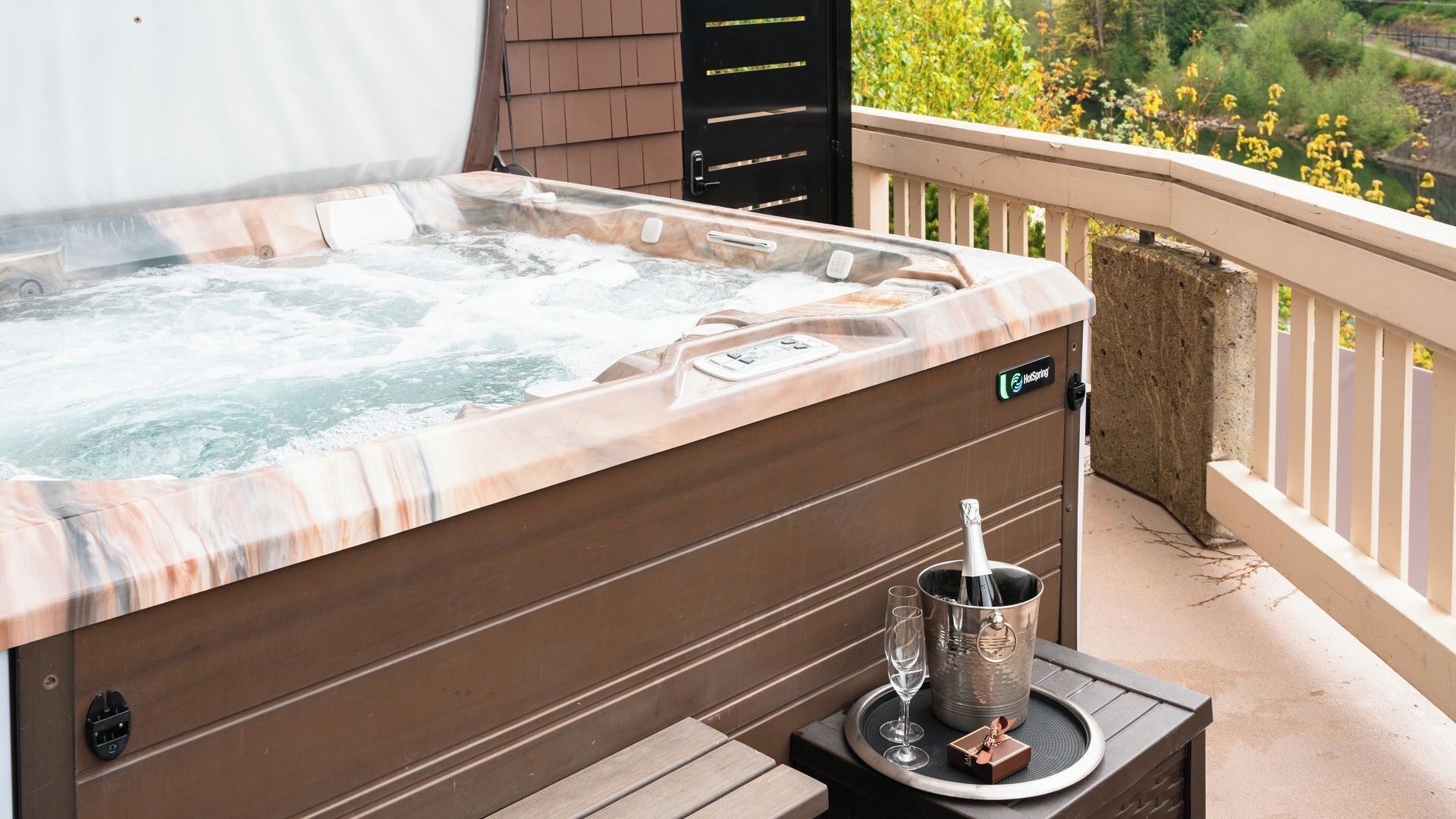 Ein Whirlpool mit Bedienfeld, ein Champagnerkühlbagger und Gläser auf einem Tisch, auf einem Balkon mit Blick auf Grün.