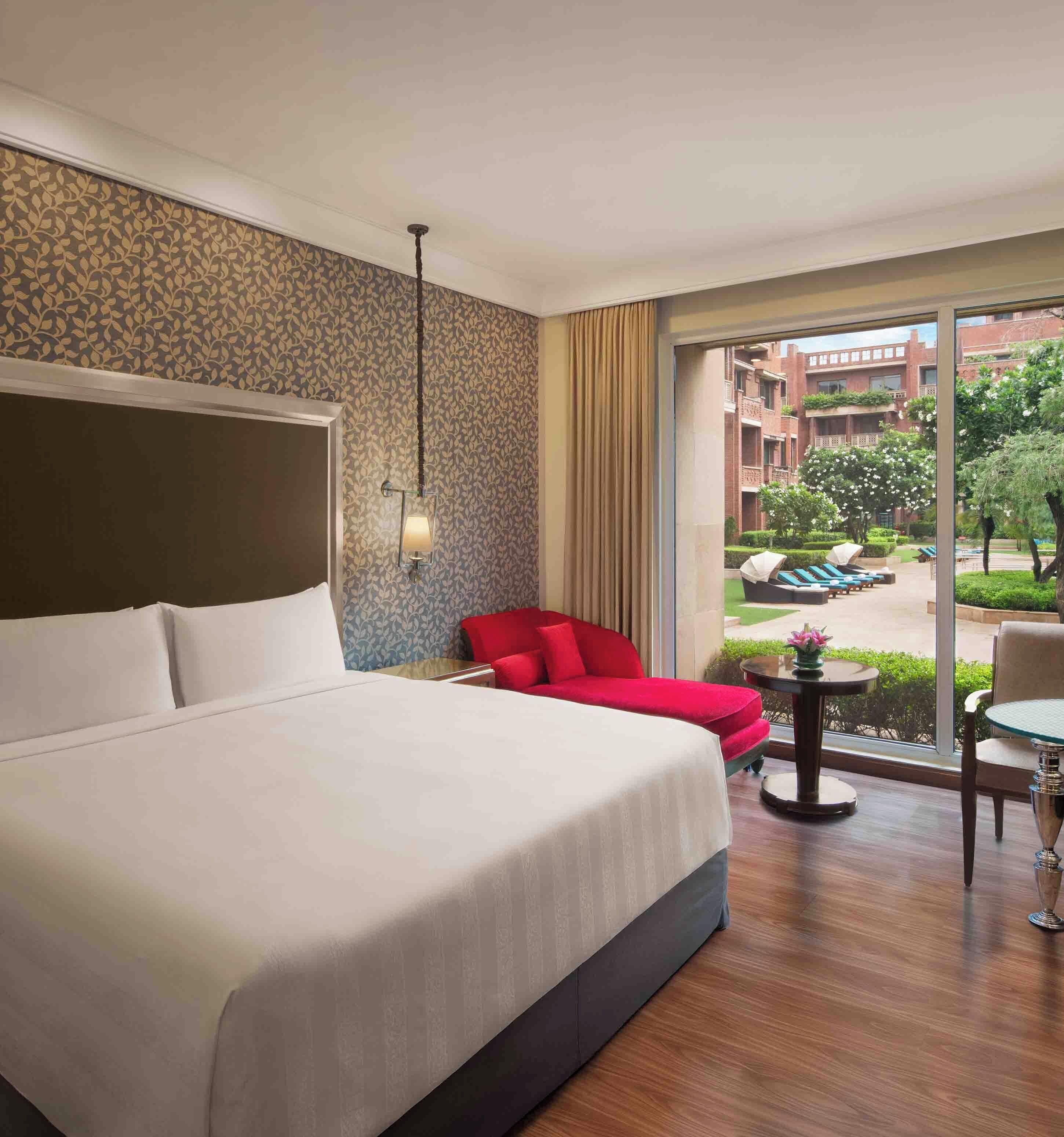 A hotel room with a large bed, a red chair, a small table, and a view of the outdoor area.