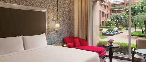 A hotel room with a large bed, a red chair, a small table, and a view of the outdoor area.