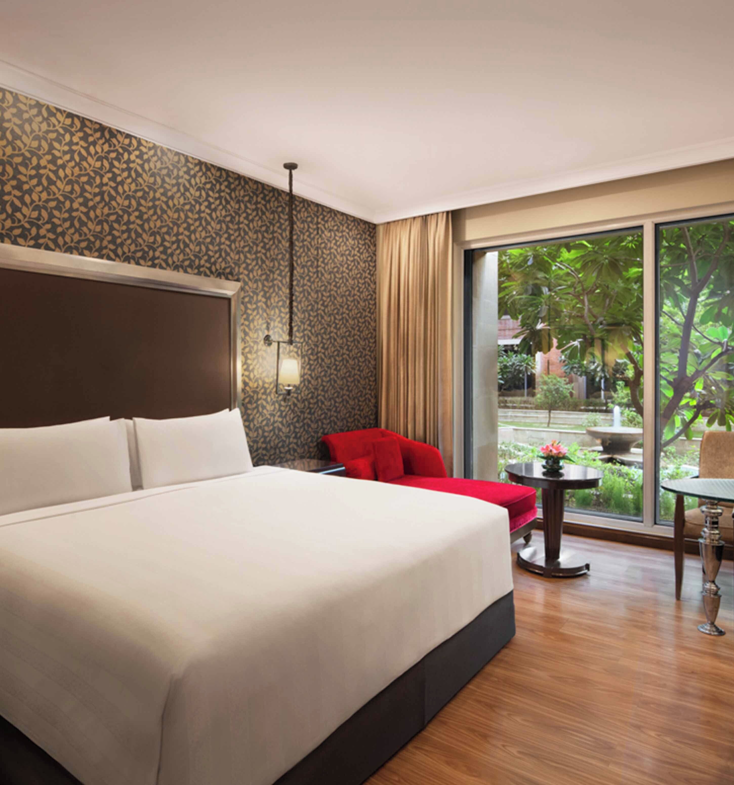 A hotel room with a large bed, a red chair, a small table, and a view of a garden through a large window.