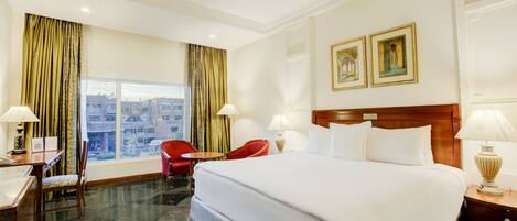 A hotel room with a large bed, two bedside lamps, a desk with a chair, a red chair, and a window with curtains.