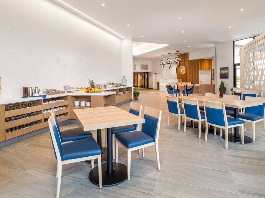 A modern dining area with wooden tables and blue cushioned chairs, a kitchen counter with utensils, and a decorative wall with circular cutouts.