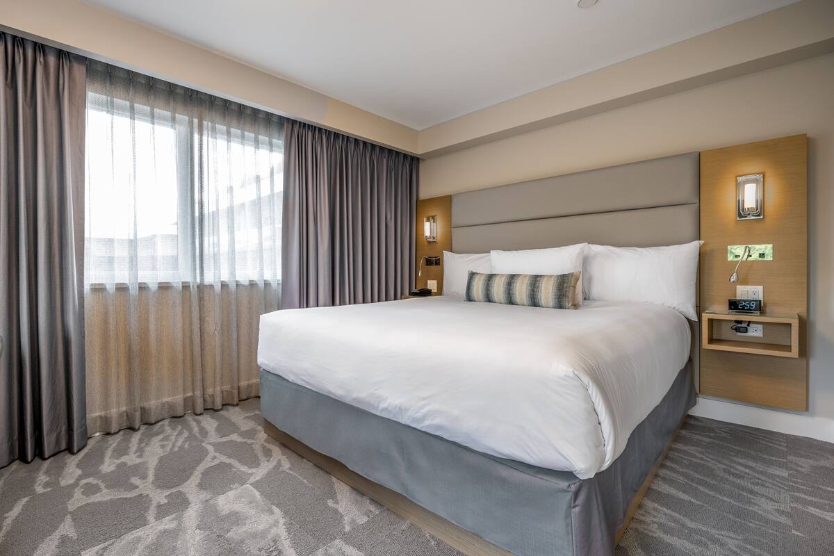 Een hotelkamer met een groot bed, een nachtkastje met klok en een raam met gordijnen.
